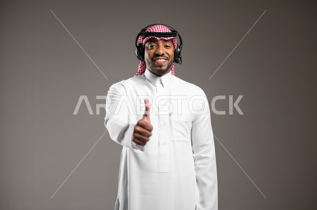 استخدام تقنية الهيدفون، تعابير الموافقة والقبول، العمل في مجال خدمة العملاء والدعم الفني، الرد على استفسارات الزبائن، بورتريه مقرب لرجل عربي خليجي سعودي يرتدي الثوب التقليدي والشماغ يضع سماعة الاتصال يرفع يده بايماءات الاعجاب، خلفية رمادية