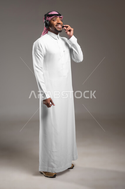 العمل في مجال خدمة العملاء والدعم الفني، الرد على استفسارات الزبائن، استخدام تقنية الهيدفون، بورتريه من الجانب لرجل عربي خليجي سعودي يرتدي الثوب التقليدي والشماغ يضع سماعة الاتصال، صورة لكامل الجسم، خلفية رمادية