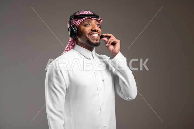 Responding to customer inquiries, working in customer service and technical support, using headphone technology, close-up side portrait of a Saudi Arabian Gulf man wearing traditional thobe and shemagh putting on a handset, gray background