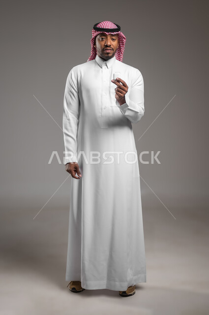 إستخدام إكسسوارات الهاتف النقال، سماعه بلوتوث حديثه متصلة بجهاز المحمول، بورتريه لرجل عربي خليجي سعودي يرتدي الزي التقليدي والشماغ يحمل بيده سماعات لاسلكيه، مفهوم التقنية والاتصالات، صورة لكامل الجسم، خلفيه رمادية