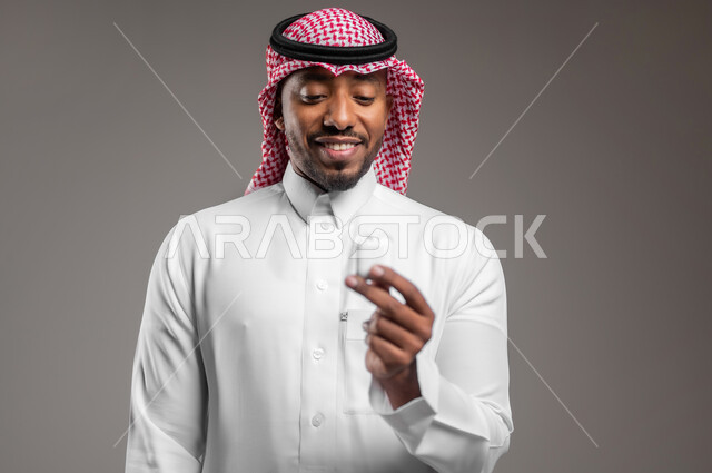 مفهوم التقنية والاتصالات، إستخدام إكسسوارات الهاتف النقال، سماعه بلوتوث حديثه متصلة بجهاز المحمول، بورتريه مقرب لرجل عربي خليجي سعودي يرتدي الزي التقليدي والشماغ يحمل بيده سماعات لاسلكيه، خلفيه رمادية
