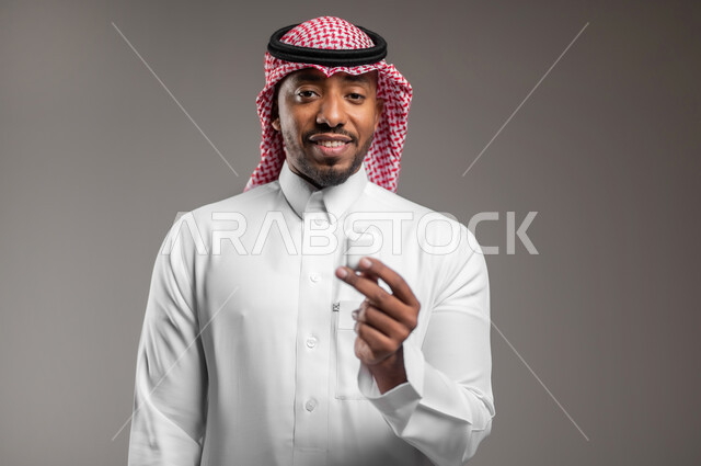 Modern Bluetooth headset connected to mobile device, close-up portrait of Saudi Arabian Gulf man wearing traditional dress and shemagh holding wireless headphones, use of mobile phone accessories, concept of technology and communication, gray background