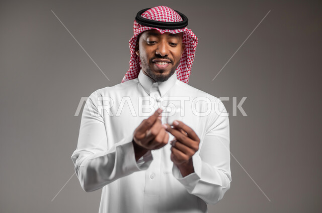 Using mobile phone accessories, modern bluetooth headset connected to mobile device, close-up portrait of a Saudi Arabian Gulf man wearing traditional dress and shemagh holding wireless headphones, technology and communication concept, gray background