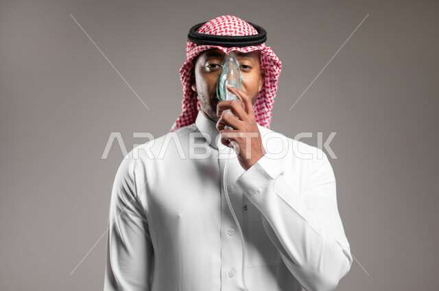 Maintaining regular airway using ventilator, close-up portrait of Saudi Arabian Gulf man wearing traditional thobe and shemagh inhaling medication for sleep apnea through oxygen mask, treatment of chronic asthma, home health care, gray background