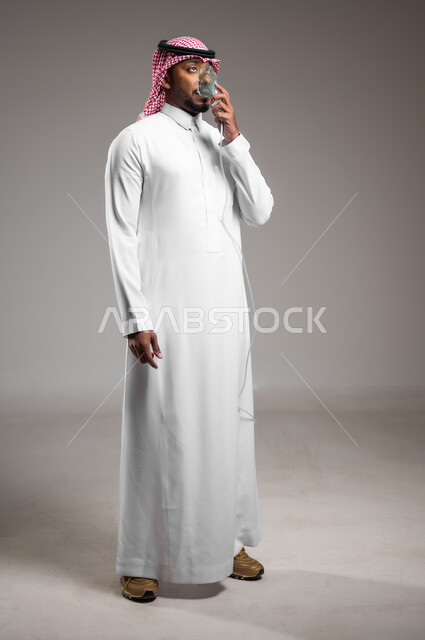 Providing health services, treating chronic asthma, maintaining a regular airway using a ventilator, portrait of a Saudi Arabian Gulf man wearing a traditional thobe and shemagh inhaling medication to treat sleep apnea through an oxygen mask, full body image, gray background