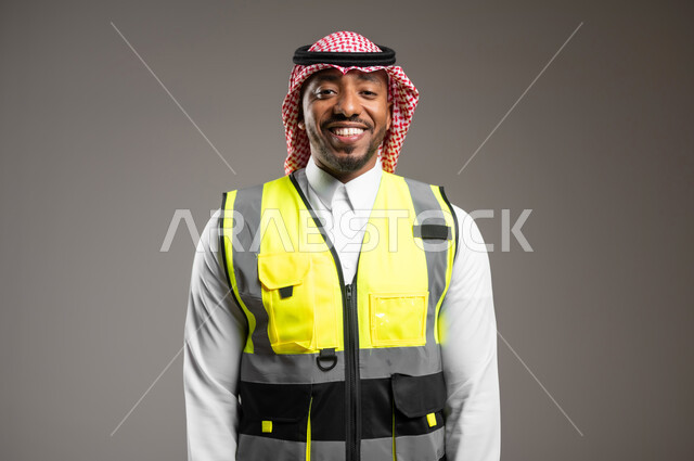 المتابعة والإشراف على خطط العمل، النظر إلى الكاميرا بايماءات السروروالسعادة، مهن و وظائف هندسية، ادارة المشاريع المعمارية الهندسية، بورتريه مقرب لمهندس عربي خليجي سعودي مبتسم يرتدي الزي التقليدي وسترة الحماية يقف باستقامة، خلفية رمادية