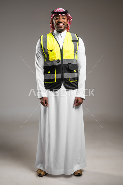 النظر إلى الكاميرا بايماءات السروروالسعادة، مهن و وظائف هندسية، ادارة المشاريع المعمارية الهندسية، المتابعة والإشراف على خطط العمل، بورتريه لمهندس عربي خليجي سعودي مبتسم يرتدي الزي التقليدي وسترة الحماية يقف باستقامة، صورة لكامل الجسم، خلفية رمادية
