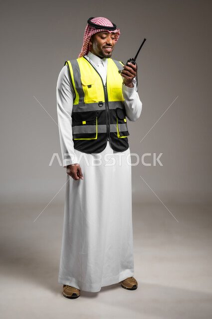 ادارة المشاريع المعمارية الهندسية، مهن و وظائف هندسية، المتابعة والإشراف على خطط العمل، بورتريه لمهندس عربي خليجي سعودي مبتسم يرتدي الزي التقليدي وسترة الحماية يقوم باستخدام اللاسلكي، صورة لكامل الجسم، خلفية رمادية