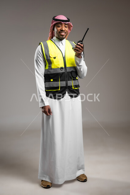 مهن و وظائف هندسية، ادارة المشاريع المعمارية الهندسية، المتابعة والإشراف على خطط العمل، بورتريه لمهندس عربي خليجي سعودي مبتسم يرتدي الزي التقليدي وسترة الحماية يقوم باستخدام اللاسلكي، صورة لكامل الجسم، خلفية رمادية