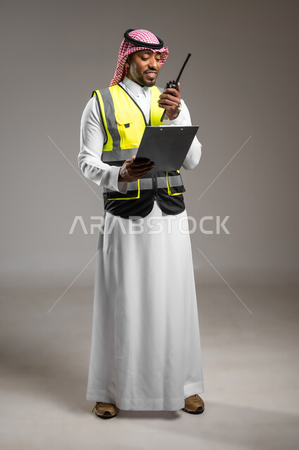 Management of Engineering Architectural Projects, Professions and Engineering Jobs, Follow -up and Supervision of Work Plans, Portrait for a smiling Saudi Arabian engineer who wears the traditional costume and a protection jacket carries with a paper holder that uses wireless, a picture of the entire body, gray background