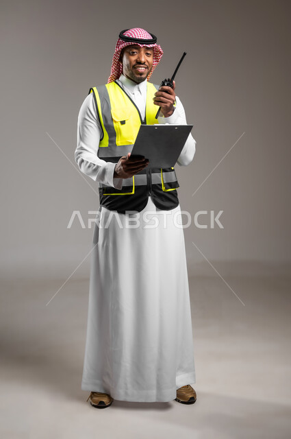 مهن و وظائف هندسية، المتابعة والإشراف على خطط العمل، بورتريه لمهندس عربي خليجي سعودي مبتسم يرتدي الزي التقليدي وسترة الحماية يحمل بيده حاملة ورق يقوم باستخدام اللاسلكي، ادارة المشاريع المعمارية الهندسية، صورة لكامل الجسم، خلفية رمادية