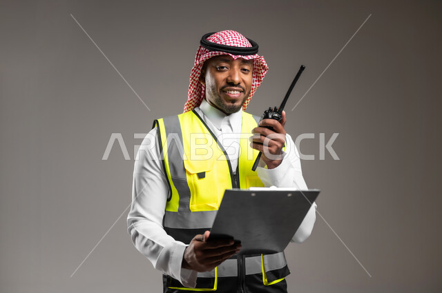 المتابعة والإشراف على خطط العمل، بورتريه مقرب لمهندس عربي خليجي سعودي مبتسم يرتدي الزي التقليدي وسترة الحماية يحمل بيده حاملة ورق يقوم باستخدام اللاسلكي، مهن و وظائف هندسية، ادارة المشاريع المعمارية الهندسية، خلفية رمادية
