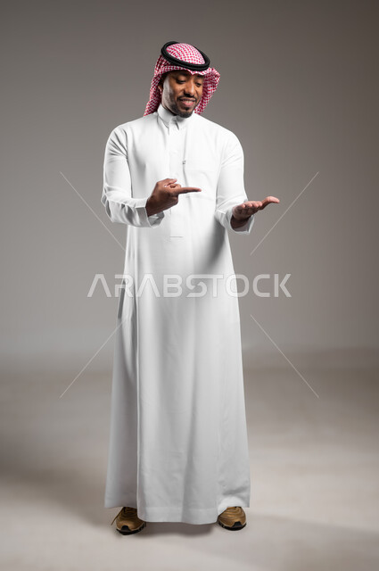 رفع شيء ما، بورتريه لرجل عربي خليجي سعودي مبتسم يرتدي الثوب التقليدي والشماغ يشير باصبع السبابة على يده، تعابير حمل عنصر هولوجرامي، صورة الجسم بالطول الكامل، خلفية رمادية