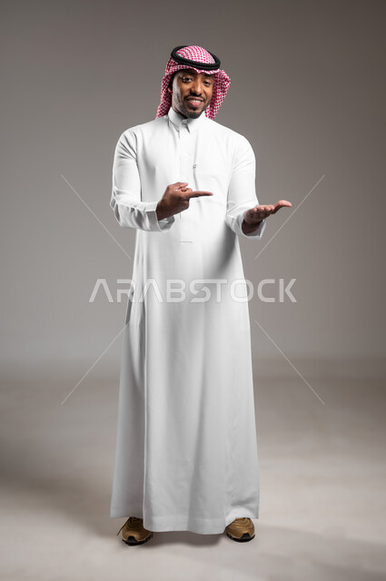 النظر إلى الكاميرا بإيماءات السرور، رفع وحمل شيء ما، بورتريه لرجل عربي خليجي سعودي مبتسم يرتدي الثوب التقليدي والشماغ يشير باصبع السبابة على يده، تعابير حمل عنصر هولوجرامي، صورة الجسم بالطول الكامل، خلفية رمادية