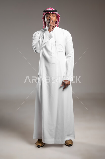 طلب السكوت والصمت، التوقف عن الكلام وخفض الصوت، الهدوء والسكون وعدم الإزعاج، بورتريه مقرب لرجل عربي خليجي سعودي يرتدي الثوب التقليدي والشماغ يضع أصبع السبابة على فمه، حركة أصبع وإشارات، صورة لكامل الجسم، خلفية رماديه