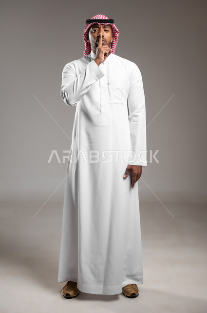 التوقف عن الكلام وخفض الصوت، الهدوء والسكون وعدم الإزعاج، بورتريه مقرب لرجل عربي خليجي سعودي يرتدي الثوب التقليدي والشماغ يضع أصبع السبابة على فمه، طلب السكوت والصمت، حركة أصبع وإشارات، صورة لكامل الجسم، خلفية رماديه