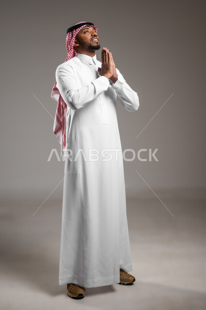 Standing with the gestures of supplication, pleading, and having, the request is to hope and humiliate the Lord of the servants, supplication to God Almighty, a portrait of a Saudi Gulf Arab man who wears the traditional garment and the shemagh raises his hands with reverence and sympathy, seeking help and desire to achieve wishes, a picture of the entire body, gray background