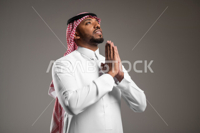 التضرع إلى الله تعالى، الوقوف بإيماءات الدعاء والتوسل والاسترحام، الطلب بترجي وتذلل من رب العباد، بورتريه مقرب لرجل عربي خليجي سعودي يرتدي الثوب التقليدي والشماغ يرفع يديه بخشوع واستعطاف، طلب العون والرغبة في تحقيق الأمنيات، خلفية رمادية
