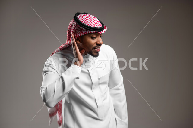 التركيز على الكلام وسماع الاسرار، الانصات وحسن الاستماع، بورتريه مقرب لرجل عربي خليجي سعودي يرتدي الثوب التقليدي والشماغ يضع يده خلف أذنه بإيماءات التنصت والتجسس، خلفية رماديه