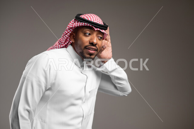 التركيز على الكلام وسماع الاسرار، الانصات وحسن الاستماع، بورتريه مقرب لرجل عربي خليجي سعودي يرتدي الثوب التقليدي والشماغ يضع يده خلف أذنه بإيماءات التنصت والتجسس، خلفية رماديه