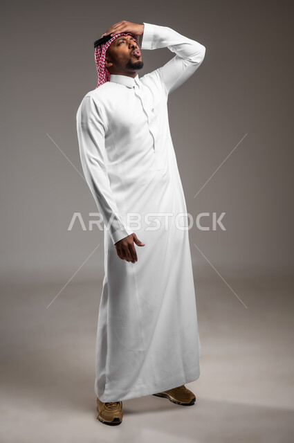 ايماءات تذكر امر ما، الشعور بالدهشة والذهول، النسيان والسهو والغفلة، بورتريه لرجل عربي خليجي سعودي يرتدي الالثوب التقليدي والشماغ يضع يده على رأسه بتعابير الصدمة، صورة لكامل الجسم، خلفية رمادية