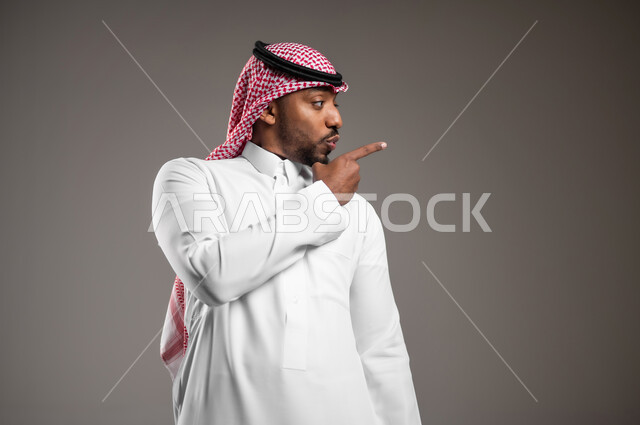 توضيح مكان ما, النظر إلى اليسار بايماءات الذهول, الإشارة إلى هدف محدد، بورتريه مقرب لشاب عربي خليجي سعودي يرتدي الثوب التقليدي والشماغ يرفع اصبع السبابة لتحديد موقع معين, خلفية رمادية
