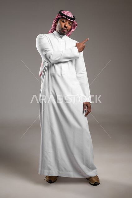 الإشارة إلى هدف محدد، النظر إلى الكاميرا بايماءات الذهول, بورتريه لشاب عربي خليجي سعودي يرتدي الثوب التقليدي والشماغ يرفع اصبع السبابة لتحديد موقع معين, توضيح مكان ما, صورة لكامل الجسم، خلفية رمادية
