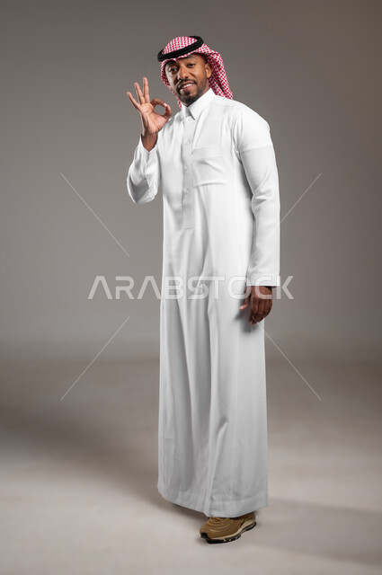 الشعور بالرضا و السعادة, حركات الاعجاب والقبول والتميز, النظر إلى الكاميرا بإشارات الموافقه، بورتريه لرجل عربي خليجي سعودي مبتسم يرتدي الثوب التقليدي والشماغ يرفع يده للأعلى بإيماءات علامة الجودة و السرور, صورة لكامل الجسم، خلفية رمادية
