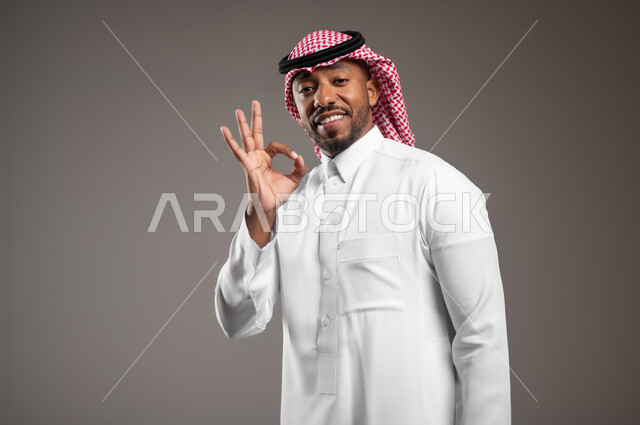 حركات الاعجاب والقبول والتميز, النظر إلى الكاميرا بإشارات الموافقه، الشعور بالرضا و السعادة, بورتريه مقرب لرجل عربي خليجي سعودي مبتسم يرتدي الثوب التقليدي والشماغ يرفع يده للأعلى بإيماءات علامة الجودة و السرور, خلفية رمادية