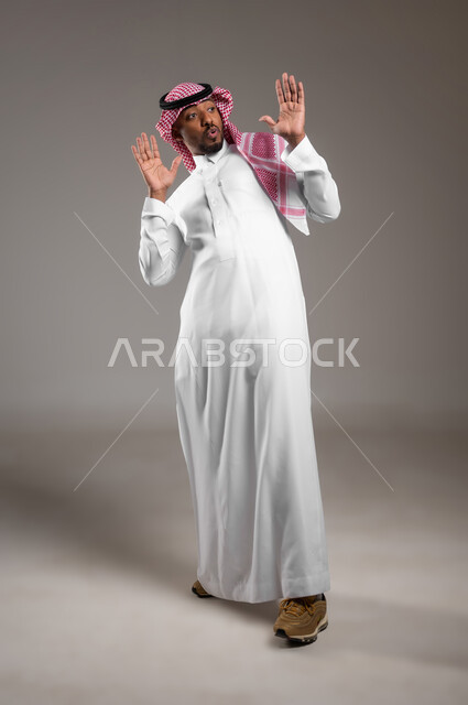 النظر إلى مكان معين بإيماءات الذعر والخوف، الشعور بالصدمة والدهشة، ايماءات التعجب والذهول، بورتريه لرجل عربي خليجي سعودي يرتدي الثوب التقليدي والشماغ يرفع يديه للأعلى بتعابير التفاجؤ لرؤية شيء ما, صورة الجسم بالكامل, خلفية رمادية