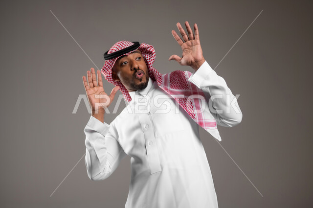 الشعور بالصدمة والدهشة، النظر إلى مكان معين بإيماءات الذعر والخوف، ايماءات التعجب والذهول، بورتريه مقرب لرجل عربي خليجي سعودي يرتدي الثوب التقليدي والشماغ يرفع يديه للأعلى بتعابير التفاجؤ لرؤية شيء ما، خلفية رمادية