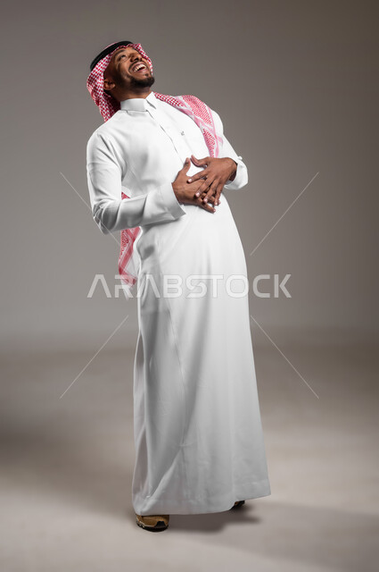 الشعور بالرضا و السعادة، تعابير البهجة والسرور الشديد، بورتريه لرجل عربي خليجي سعودي يرتدي الثوب التقليدي والشماغ يضع يده على بطنة بإيماءات الضحك الهستيرية، صورة لكامل الجسم، خلفية رمادية