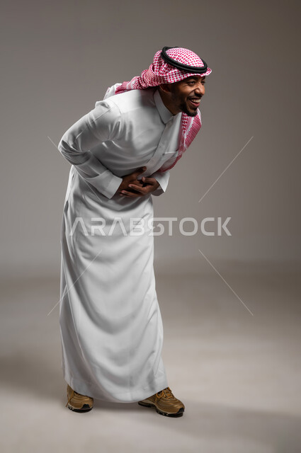 تعابير البهجة والسرور الشديد، الشعور بالرضا و السعادة، بورتريه لرجل عربي خليجي سعودي يرتدي الثوب التقليدي والشماغ يضع يده على بطنة بإيماءات الضحك الهستيرية، صورة لكامل الجسم، خلفية رمادية