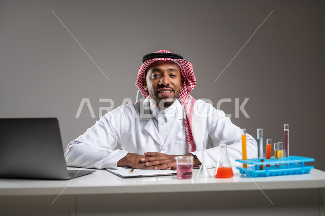 Studies and experiments in an advanced practical environment, acquire knowledge and experience with accuracy and concentration gestures, development and research and conduct chemical reactions, a close portrait of a Saudi Gulf Arab man who wears traditional costume and prevention glasses sitting in front of the mobile computer performs a scientific experience, gray background