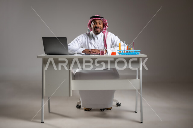 Acquiring knowledge with the gestures of focus, studies and experiments in an advanced practical environment, development, research and conducting chemical reactions, portrait for a Saudi Gulf Arab man who wears traditional costume and prevention glasses sitting in front of the mobile computer performs a scientific experience, a picture of the entire body, gray background