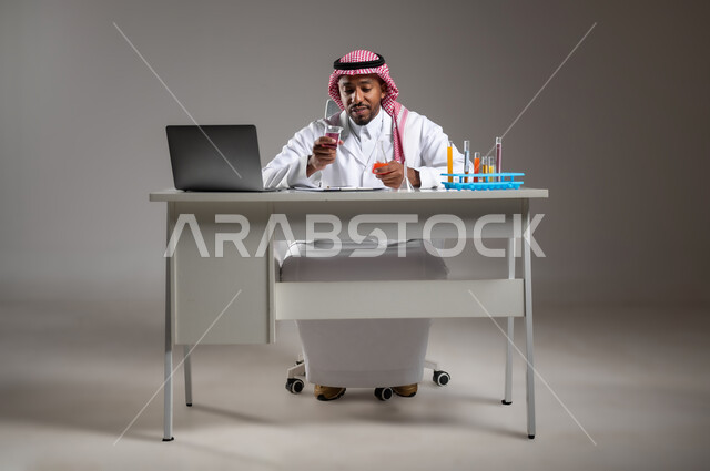 البحث وإجراء التفاعلات الكيميائية, اكتساب المعرفة بإيماءات التركيز، الدراسات والتجارب في بيئة عملية متطورة, بورتريه لرجل عربي خليجي سعودي يرتدي الزي التقليدي و نظارة الوقاية يجلس امام الحاسوب المحمول يقوم بتجربة علمية, صورة لكامل الجسم، خلفية رمادية