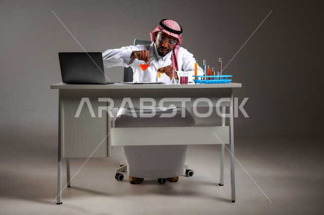 التطوير والبحث وإجراء التفاعلات الكيميائية, اكتساب المعرفة بإيماءات التركيز، الدراسات والتجارب في بيئة عملية متطورة, بورتريه لرجل عربي خليجي سعودي يرتدي الزي التقليدي و نظارة الوقاية يجلس امام الحاسوب المحمول يقوم بتجربة علمية, صورة لكامل الجسم، خلفية رمادية