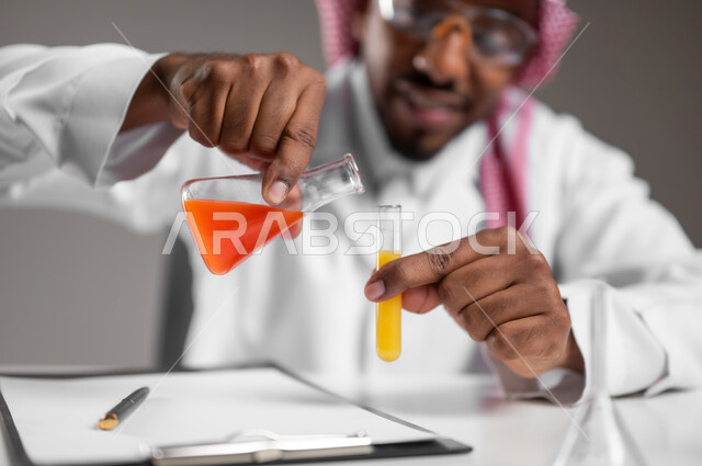 Acquiring knowledge and experience with the gestures of accuracy and concentration, studies and experiences in an advanced practical environment, development and research and conducting chemical reactions, a close portrait of a Saudi Gulf Arab man wearing traditional costume and prevention glasses sitting in front of the mobile computer performs a scientific experience, gray background