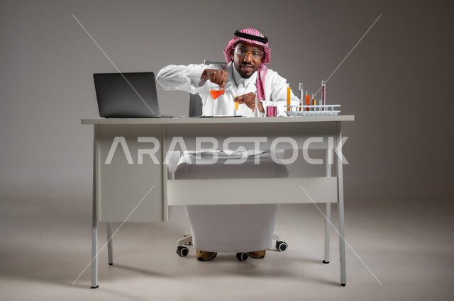 اكتساب المعرفة بإيماءات التركيز، الدراسات والتجارب في بيئة عملية متطورة, التطوير والبحث وإجراء التفاعلات الكيميائية, بورتريه لرجل عربي خليجي سعودي يرتدي الزي التقليدي و نظارة الوقاية يجلس امام الحاسوب المحمول يقوم بتجربة علمية, صورة لكامل الجسم، خلفية رمادية