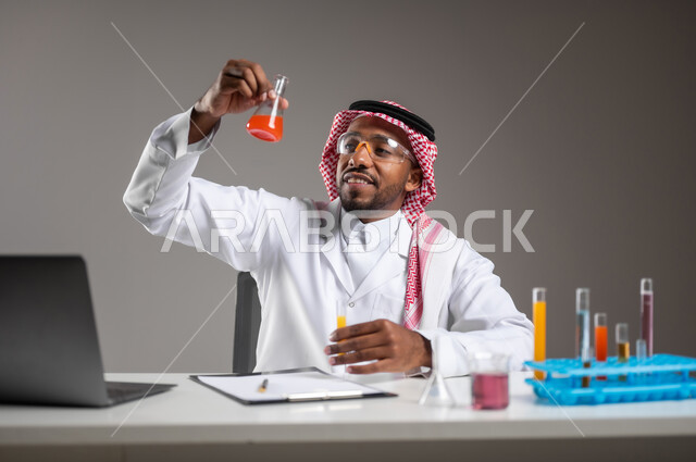 Acquiring knowledge and experience with the gestures of accuracy and concentration, studies and experiences in an advanced practical environment, development and research and conducting chemical reactions, a close portrait of a Saudi Gulf Arab man wearing traditional costume and prevention glasses sitting in front of the mobile computer performs a scientific experience, gray background