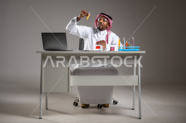التطوير والبحث وإجراء التفاعلات الكيميائية, اكتساب المعرفة بإيماءات التركيز، الدراسات والتجارب في بيئة عملية متطورة, بورتريه لرجل عربي خليجي سعودي يرتدي الزي التقليدي و نظارة الوقاية يجلس امام الحاسوب المحمول يقوم بتجربة علمية, صورة لكامل الجسم، خلفية رمادية