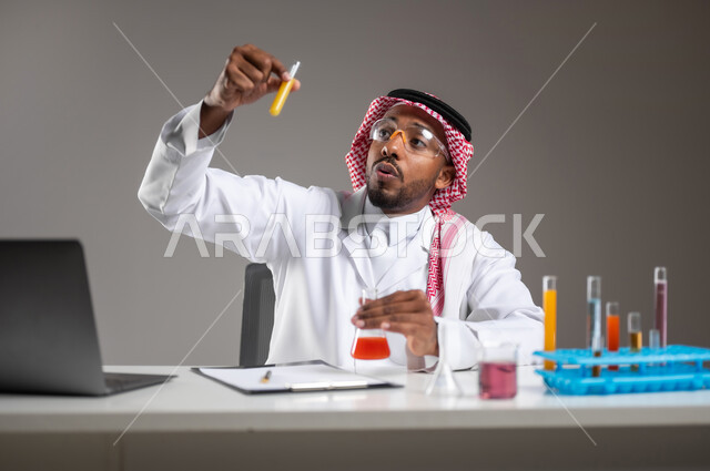 الدراسات والتجارب في بيئة عملية متطورة, اكتساب المعرفة والخبرة بإيماءات الدقة والتركيز، التطوير والبحث وإجراء التفاعلات الكيميائية, بورتريه مقرب لرجل عربي خليجي سعودي يرتدي الزي التقليدي و نظارة الوقاية يجلس امام الحاسوب المحمول يقوم بتجربة علمية, خلفية رمادية