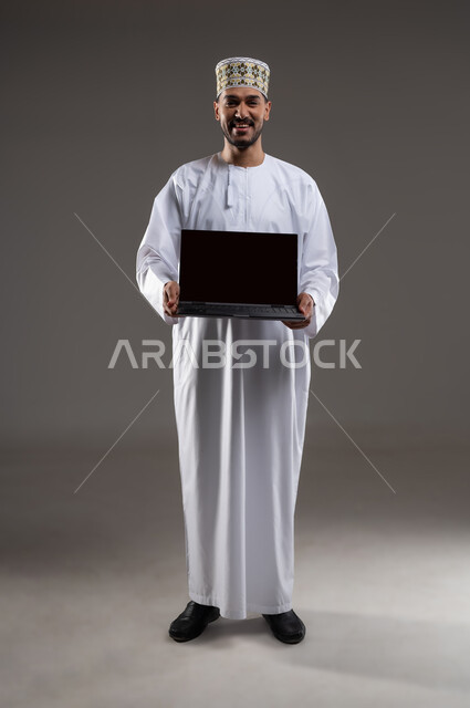 مفهوم استخدام التقنيات الحديثة والأجهزة المتطورة ، بورتريه بالطول الكامل لشاب عربي خليجي عماني يرتدي الدشداشة والكمه يحمل بيده جهاز الحاسب المحمول تظهر عليه علامات الفرح والسرور، انجاز المهام والعمل عن بعد، خلفية رماديه