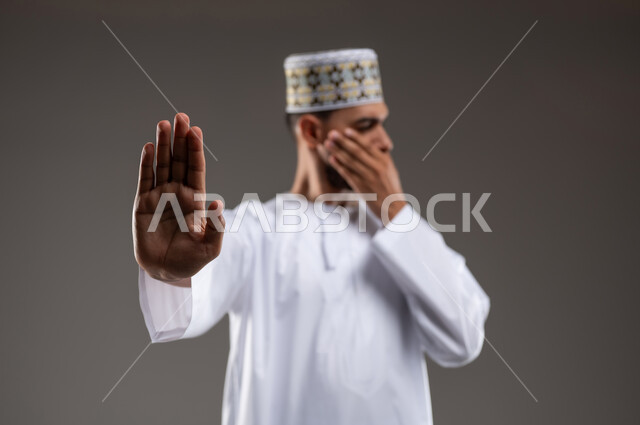 Command to stop doing something, expressions of refusal and abstention, close-up portrait of a young Gulf Arab Omani man wearing a dishdasha and a turban, gesturing with his hand and face with gestures of prohibition and feeling angry and upset, gray background