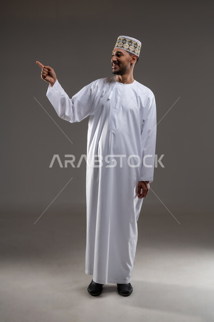 الإشارة والتوجيه الى مكان ما، بورتريه لرجل عربي خليجي عماني يرتدي الدشداشة والكمه يقف مبتسما ويشير بيده الى مكان ما، تحديد موقع  او هدف معين، حركات واشارات، خلفيه رماديه