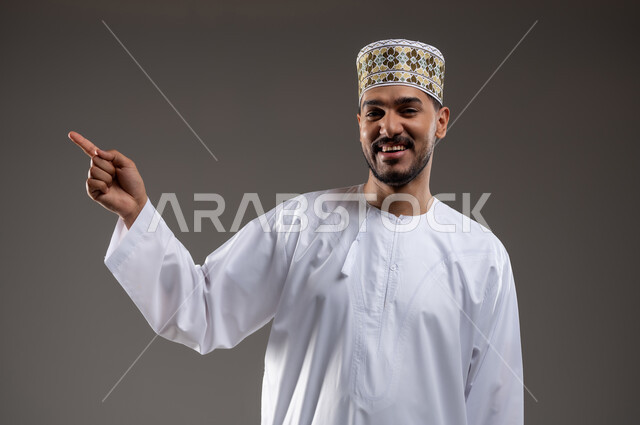 الإشارة والتوجيه الى مكان ما، بورتريه لرجل عربي خليجي عماني يرتدي الدشداشة والكمه يقف مبتسما ويشير بيده الى مكان ما، تحديد موقع  او هدف معين، حركات واشارات، خلفيه رماديه