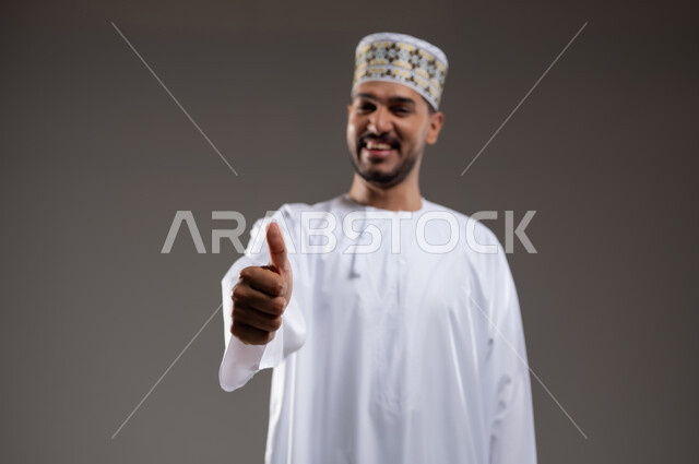 ايماءات تدل على الموافقة والقبول، بورتريه لرجل عربي خليجي عماني يرتدي الدشداشة والكمه يشير باصبع الابهام للاعلى تظهر عليه تعابير الفرح والسرور، الرضى وعدم الرفض، خلفية رماديه
