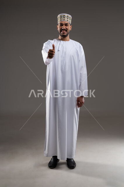 ايماءات تدل على الموافقة والقبول، بورتريه لرجل عربي خليجي عماني يرتدي الدشداشة والكمه يشير باصبع الابهام للاعلى تظهر عليه تعابير الفرح والسرور، الرضى وعدم الرفض، خلفية رماديه