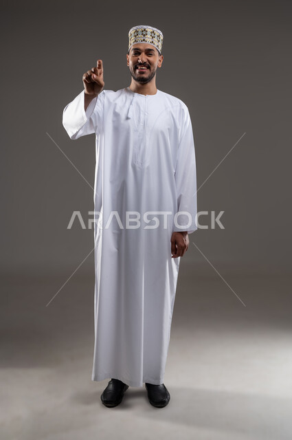 الإشارة والتوجيه الى مكان ما، بورتريه لرجل عربي خليجي عماني يرتدي الدشداشة والكمه يقف مبتسما ويشير بيده الى مكان ما، تحديد موقع  او هدف معين، حركات واشارات، خلفيه رماديه
