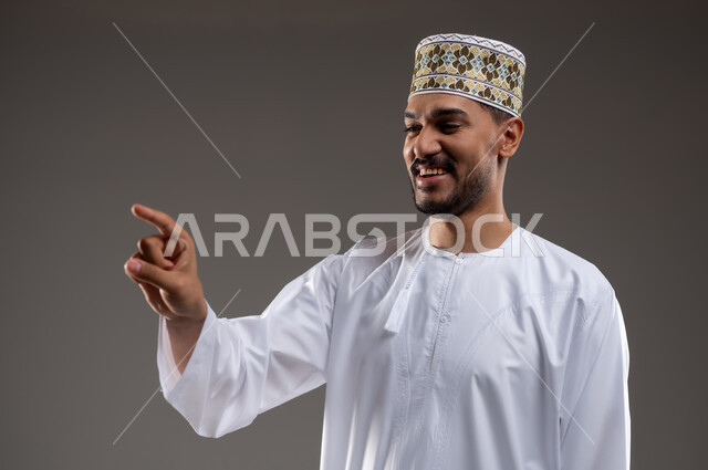 الإشارة والتوجيه الى مكان ما، بورتريه لرجل عربي خليجي عماني يرتدي الدشداشة والكمه يقف مبتسما ويشير بيده الى مكان ما، تحديد موقع  او هدف معين، حركات واشارات، خلفيه رماديه